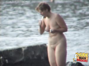 Зрелая нудистка рано утром чистит зубы на пляже